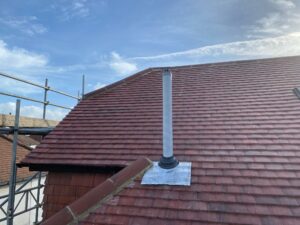 A roof with red tiles features a white vent pipe and metal flashing, under a blue sky with scattered clouds and scaffolding in the background.