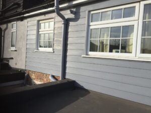 Side view of a house featuring gray siding, two white windows, and a downpipe, with a flat surface in the foreground.