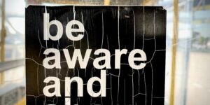 A cracked and weathered black sign with bold white text that reads "be aware and share," displayed behind scratched glass in an industrial setting.